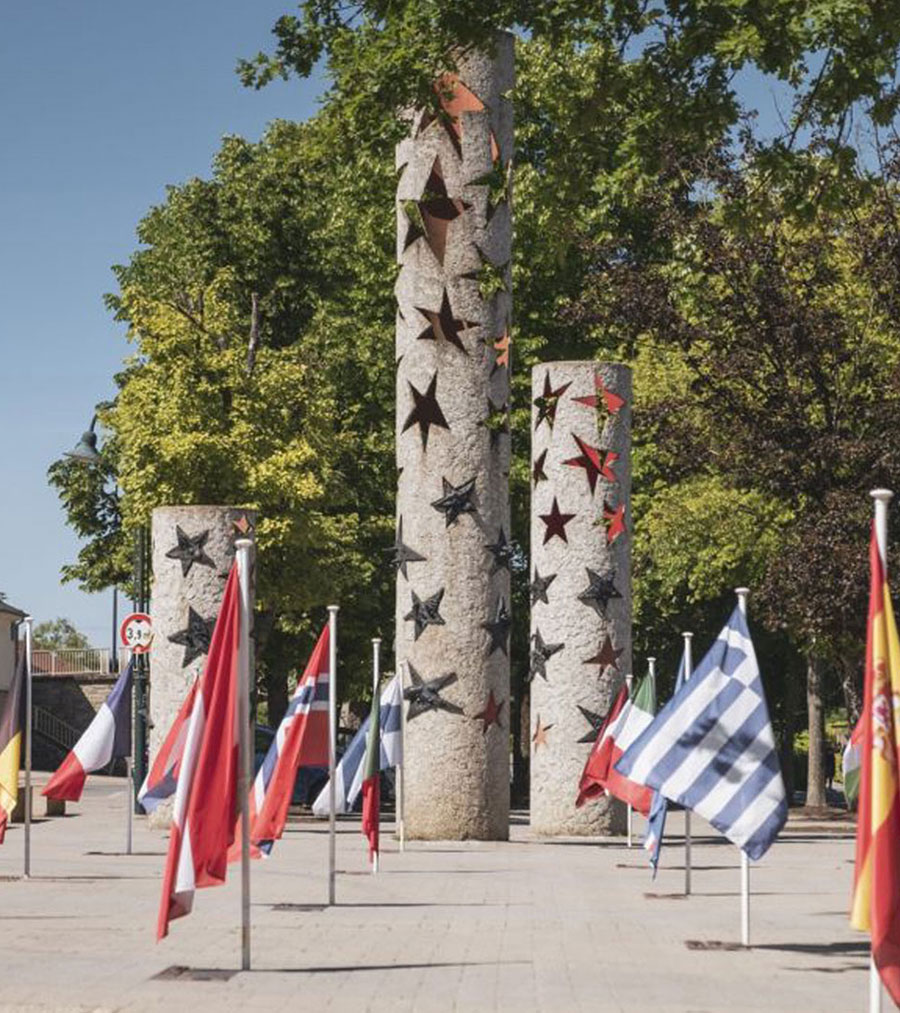 Three cylindrical stone columns adorned with colorful stars stand amid flagpoles displaying international flags. The scene is serene and bright.