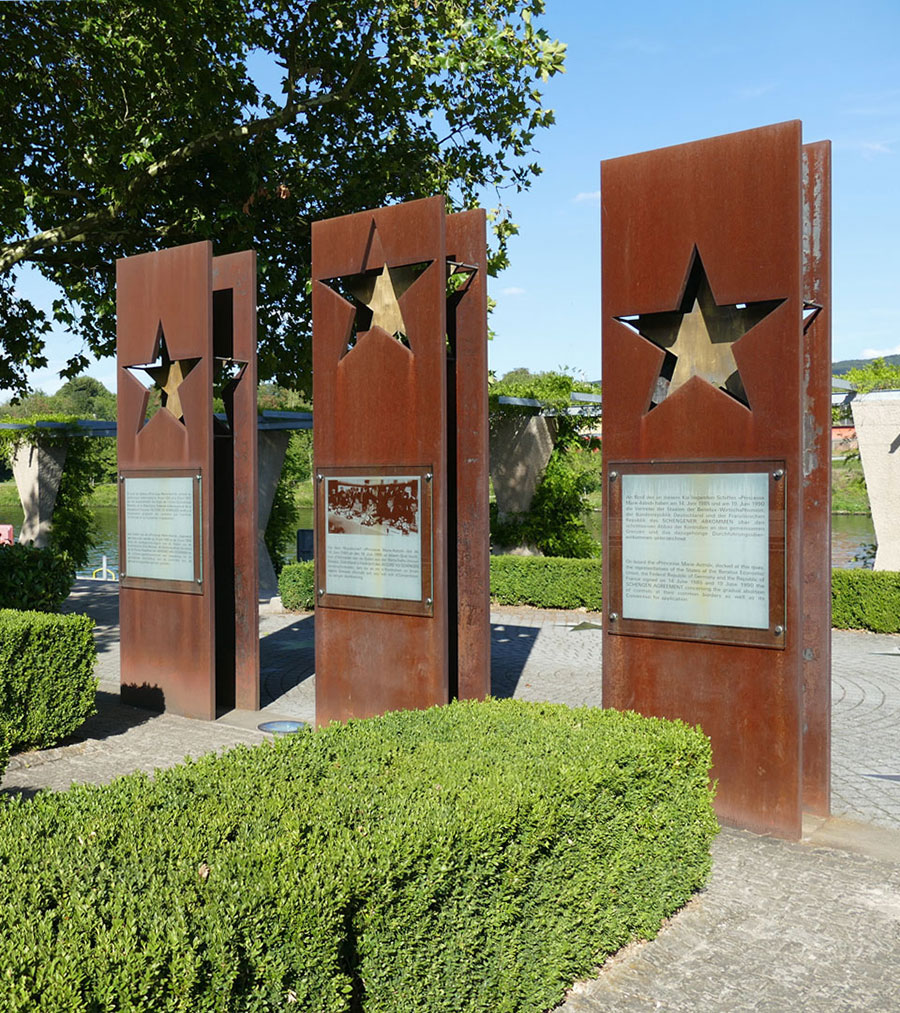 Three rust-colored metal panels with star cutouts stand upright in a garden. Each displays a glass-encased plaque with text. Green hedges surround the area.